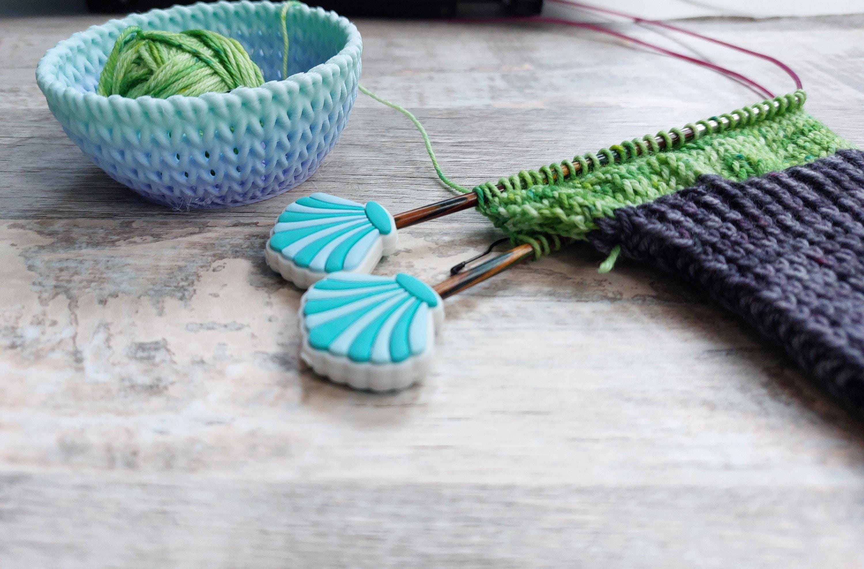 May include: A light blue bowl contains a ball of green yarn. Two seashell stitch markers sit near knitting needles with green and purple knitted fabric. The needles are inserted into the knitted fabric. The scene is on a wooden surface.